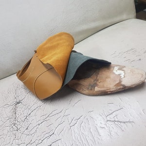 Puede incluir: Una parte superior de zapato de cuero marr&oacute;n con una horma de zapato de madera parcialmente construida. Un trozo de cuero marr&oacute;n y verde azulado est&aacute; drapeado sobre el zapato. El fondo es una superficie blanca con grietas.