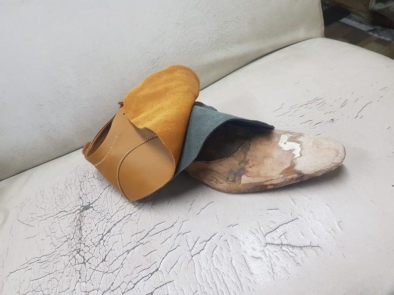 Puede incluir: Una parte superior de zapato de cuero marr&oacute;n con una horma de zapato de madera parcialmente construida. Un trozo de cuero marr&oacute;n y verde azulado est&aacute; drapeado sobre el zapato. El fondo es una superficie blanca con grietas.