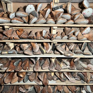 May include: A collection of wooden shoe lasts, in various sizes and shapes, neatly arranged on wooden shelves. The lasts are primarily brown, with some showing signs of wear and age. The image suggests a workshop or storage area for shoemaking supplies.