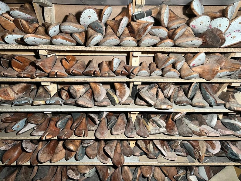 May include: A collection of wooden shoe lasts, in various sizes and shapes, neatly arranged on wooden shelves. The lasts are primarily brown, with some showing signs of wear and age. The image suggests a workshop or storage area for shoemaking supplies.