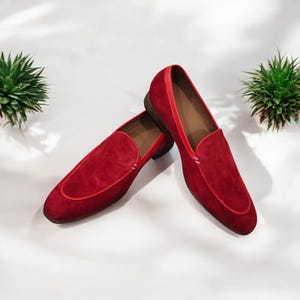 Könnte beinhalten: Ein Paar rote Wildleder-Loafer mit Lederausstattung. Die Schuhe haben ein klassisches Slip-on-Design mit runder Spitze und niedrigem Absatz. Die Innenseite ist hellbraun, vor weißem Hintergrund.