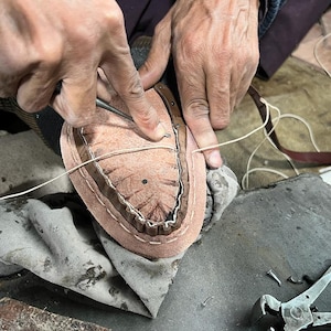 Puede incluir: Primer plano del proceso de fabricaci&oacute;n de zapatos. Las manos de un zapatero cosen la suela de un zapato. La suela del zapato es de color marr&oacute;n claro, con un borde marr&oacute;n m&aacute;s oscuro. Hilo blanco y otras herramientas tambi&eacute;n visibles.