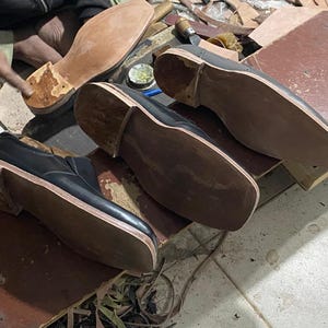 May include: A workshop scene featuring partially constructed black leather shoes with light brown soles. The shoes are in various stages of assembly, with wooden lasts and tools visible. The focus is on the craftsmanship of shoemaking.