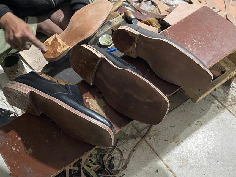 May include: A workshop scene featuring partially constructed black leather shoes with light brown soles. The shoes are in various stages of assembly, with wooden lasts and tools visible. The focus is on the craftsmanship of shoemaking.