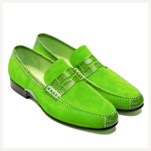 May include: A pair of bright green suede loafers with a crocodile-textured leather strap across the top. The shoes have white stitching and a black sole. The overall style is classic and fashionable.