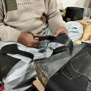 May include: A person cutting black leather with scissors. The leather pieces have white stitching and are likely for a shoe or boot. The person is wearing a beige sweater and sitting on a light blue surface.