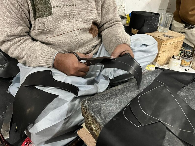May include: A person cutting black leather with scissors. The leather pieces have white stitching and are likely for a shoe or boot. The person is wearing a beige sweater and sitting on a light blue surface.