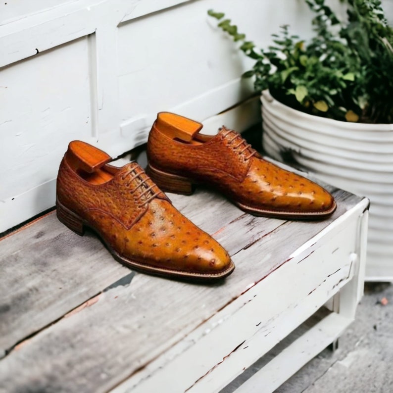 Puede incluir: Un par de zapatos de vestir de cuero marr&oacute;n con un patr&oacute;n de avestruz texturizado. Los zapatos tienen un dise&ntilde;o cl&aacute;sico con cordones y est&aacute;n colocados sobre una superficie de madera envejecida. Hormas de zapato insertadas en cada zapato.