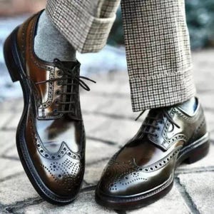 May include: A pair of polished brown leather brogue shoes with intricate perforations and a glossy finish. The shoes feature a classic lace-up design and are worn with gray socks and plaid trousers.