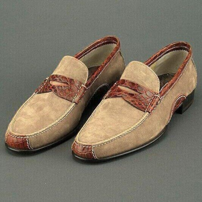 Puede incluir: Un par de mocasines penny de ante color beige con detalles de cuero marr&oacute;n con textura de cocodrilo. Los zapatos presentan un dise&ntilde;o cl&aacute;sico con punta redonda, una correa de cuero en la pala y suela cosida.