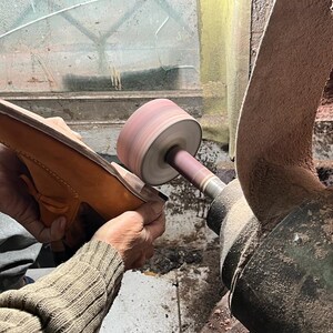 May include: A shoemaker sands the sole of a tan leather loafer with a sanding machine. The shoe is held in place by the shoemaker's hands. The machine has a rotating sanding wheel and a large, curved sanding belt.