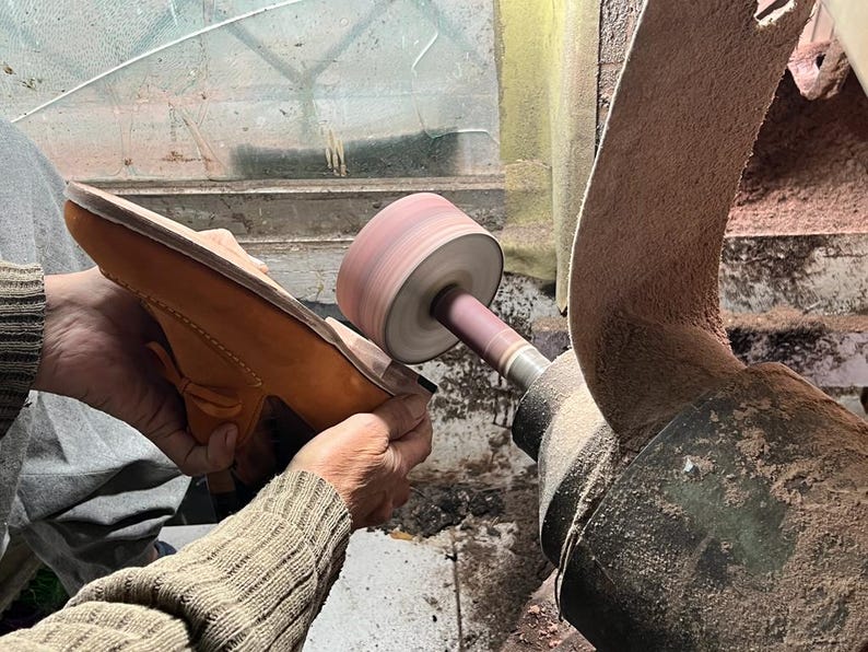 May include: A shoemaker sands the sole of a tan leather loafer with a sanding machine. The shoe is held in place by the shoemaker's hands. The machine has a rotating sanding wheel and a large, curved sanding belt.