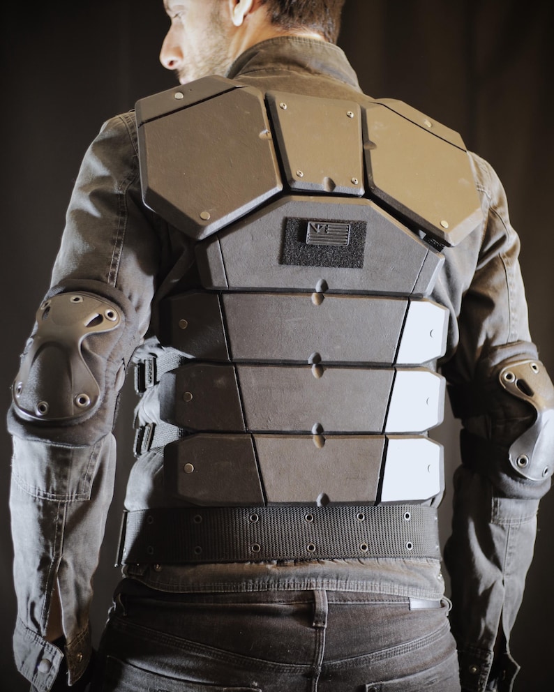 May include: A black tactical vest with multiple adjustable straps and a black patch with a white flag design. The vest has a segmented, armored front panel.