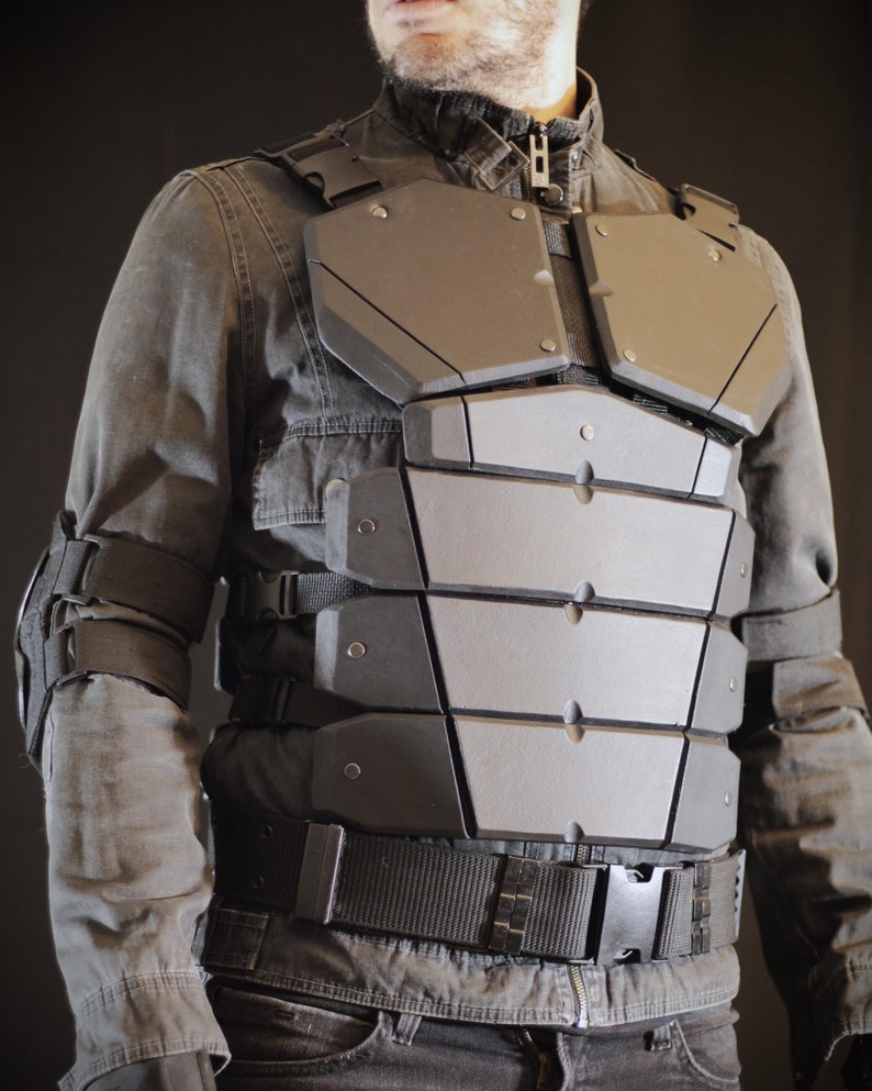 May include: A black tactical vest with multiple straps and buckles. The vest has a segmented, armored chest plate with a geometric design.
