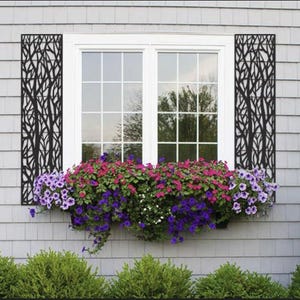 May include: A white window with a flower box filled with purple, pink, and white petunias. Black metal tree-shaped decorative panels flank the window. The house exterior is gray with green bushes below.