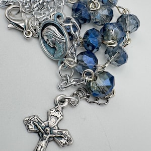 Op de afbeelding: Een rozenkrans met gefacetteerde donkerblauwe kristallen kralen verbonden door een zilverkleurige metalen ketting. Het sieraad bevat een zilverkleurig kruisbeeld en een ronde medaille met het profiel van een figuur, afgewerkt met een karabijnsluiting.