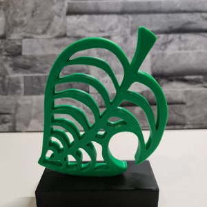 May include: A green 3D printed figurine of a leaf with a hole in the middle, on a black base. The leaf is a popular symbol from the video game Animal Crossing.