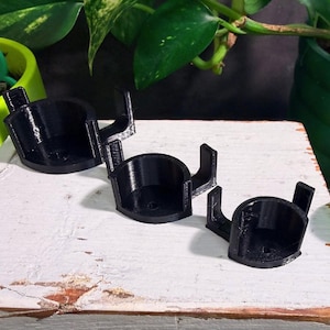 May include: Three black, 3D-printed plant pot holders. Each holder has a circular base with two curved arms extending upwards. The holders are arranged on a white, weathered wooden surface, with green plants in the background.