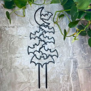 May include: Black metal bat and moon-shaped garden stake. The stake features a crescent moon with a bat silhouette and a stack of bat silhouettes. The stake has two prongs for easy placement in soil. A decorative accent for Halloween or a gothic garden.