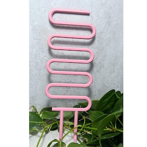 Könnte beinhalten: Ein rosa Pflanzenstützstab mit wellenförmigem Design. Der Stab besteht aus einem festen Material und hat unten zwei Zinken zum Einsetzen in den Boden. Der Stab wird vor grünen Blättern gezeigt.