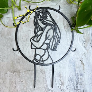 May include: Black metal cake topper featuring a silhouette of a woman with long hair, arms crossed, within a circular frame. A yellow constellation is above the frame. The topper has two stakes for insertion into a cake.