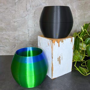 May include: Two decorative planters. One is black and sits atop a white wooden block. The other is green and blue. Both have a rounded, oval shape, suitable for small plants or succulents.