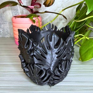 May include: A black planter with a leaf design. The planter is made of a dark material and features a detailed pattern of leaves around the exterior. The planter is designed to hold plants and is suitable for indoor or outdoor use.
