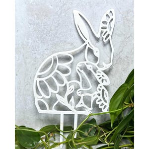May include: White metal cutout of a rabbit with a smaller rabbit inside. The rabbit is decorated with a floral pattern. The cutout is on a white background.