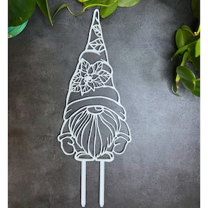 May include: A white, laser-cut garden gnome stake. The gnome has a pointed hat with leaf details, a floral design, and a long beard. Two stakes extend from the bottom for easy placement in soil. The design is intricate and detailed.