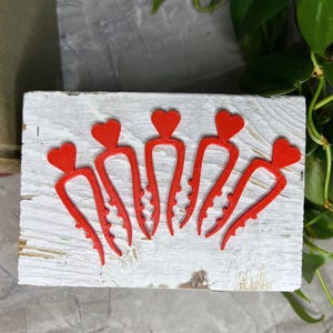 May include: Five red heart-shaped hair clips arranged on a white, weathered wooden block. Each clip has a heart at the top and a curved, toothed design. The background includes greenery and a gray surface.