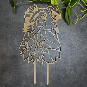 May include: A wooden cake topper featuring a detailed silhouette of a dog's head, with intricate floral and leaf-like patterns. The topper has two slender stakes for insertion into a cake. The design is in a natural wood tone.