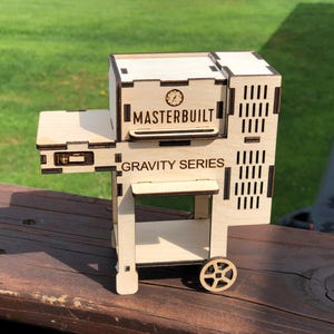 May include: A wooden model of a Masterbuilt Gravity Series grill. The model is made of laser-cut wood and features a detailed design. The grill has a removable lid, a side shelf, and a bottom shelf. The model is painted in a natural wood finish.