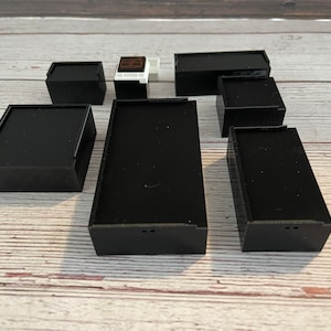 May include: Eight black square-shaped boxes of various sizes, some with small holes on the bottom, on a light brown wooden surface.