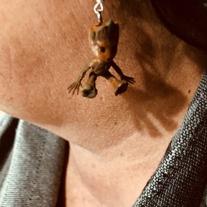 May include: Silver dangle earrings featuring a small, brown, wooden figurine of a character from the Guardians of the Galaxy movie. The figurine is sitting with its arms outstretched.