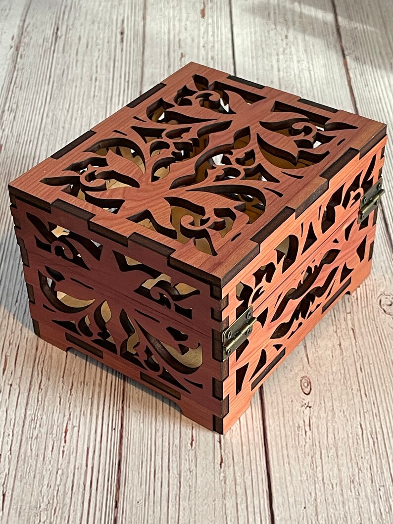 May include: A wooden box with a hinged lid and intricate floral cutouts. The box is made of a reddish-brown wood and has a natural wood grain finish.