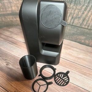 May include: A black 3D printed object with a round, flat top and a cylindrical base. The object has a small, round, flat piece attached to the top. There are four smaller 3D printed objects, including a round ring, a ring with three prongs, and a round piece with a grid pattern.