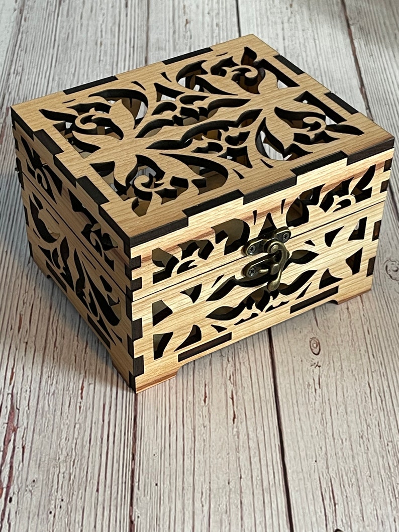 May include: A wooden box with a decorative, intricate floral design cut out of the lid. The box has a brown latch and is made of light wood.