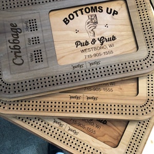 May include: Three wooden cribbage boards with a laser-engraved design. The top board has a sign that reads "Bottoms Up Pub & Grub Westboro, WI 715-905-1555".