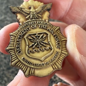 Könnte beinhalten: Ein goldfarbenes Metallabzeichen der Security Police des US-Luftwaffenministeriums. Das Abzeichen zeigt einen Adler oben und die Worte "Department of the Air Force" und "United States of America" am Rand.