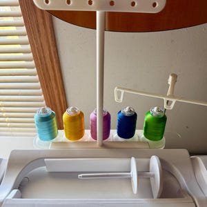 Puede incluir: Una máquina de coser blanca con un soporte para carretes y un devanador de canillas. El soporte para carretes tiene seis carretes de hilo en los colores turquesa, amarillo, rosa, azul, verde y blanco.