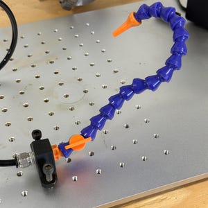 Puede incluir: Una manguera flexible de refrigerante azul con una boquilla y accesorios naranjas. La manguera está sujeta a un soporte negro y montada en una placa metálica con agujeros espaciados uniformemente. La manguera está diseñada para dirigir fluidos.