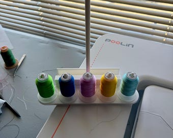 Pode incluir: Um suporte de fio de plástico branco com seis carreteis de fio. Os carreteis são verdes, azuis, rosa, amarelos e azul claro. O suporte é etiquetado como "POOLin".
