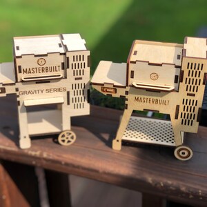 May include: Two miniature wooden models of Masterbuilt gravity series grills. The grills are made of light brown wood and have a black grill grate. The grills are on a wooden surface.