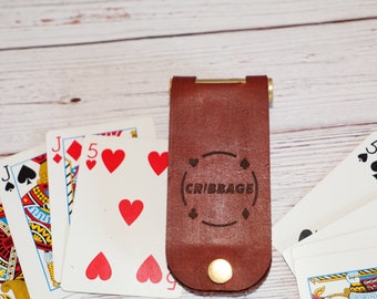 以下が含まれることがあります： ゴールドのアクセントが付いた茶色の革製のクリbbageカードホルダーで、前面に「CRIBBAGE」という文字が刻印されています。ホルダーはトランプで囲まれています。