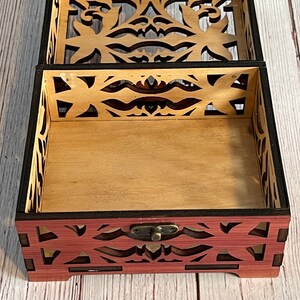 May include: A wooden box with a decorative, intricate cut-out design. The box is open, revealing a light brown interior. The box has a dark brown border and a brass latch.