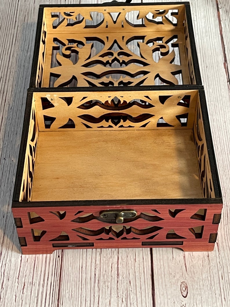May include: A wooden box with a decorative, intricate cut-out design. The box is open, revealing a light brown interior. The box has a dark brown border and a brass latch.