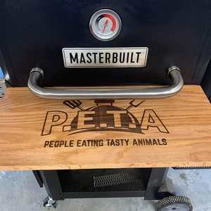 Könnte beinhalten: Ein schwarzer Masterbuilt-Smoker mit einem Holzbrett auf der Oberseite. Das Brett hat ein eingebranntes Design, auf dem "P.E.T.A. People Eating Tasty Animals" steht.