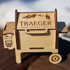 May include: A miniature wooden model of a Traeger wood pellet grill. The model is light brown and features the Traeger logo and the words "Wood Pellet Grills".
