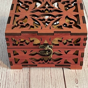 May include: A wooden box with a decorative, intricate, laser-cut design. The box has a hinged lid and a brass latch.