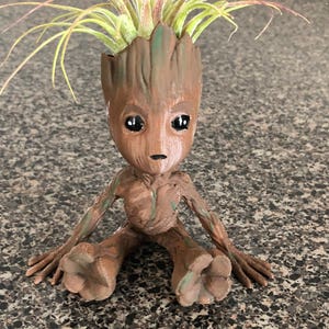 Puede incluir: Un macetero de cerámica marrón y verde con forma de personaje sentado de la película Guardianes de la Galaxia. El macetero tiene una planta de aire verde que crece en la parte superior.